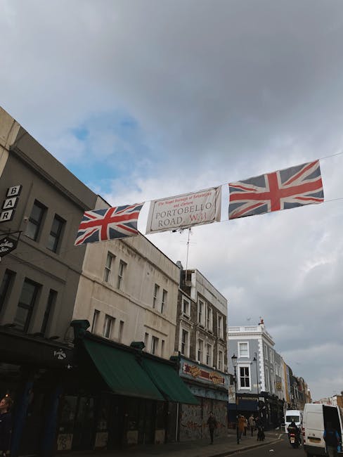 A street scene on Portobello Road in Notting Hill, with a banner displaying the British flag and the street name, hanging above the pavement. The sky is partly cloudy with patches of blue visible. On 