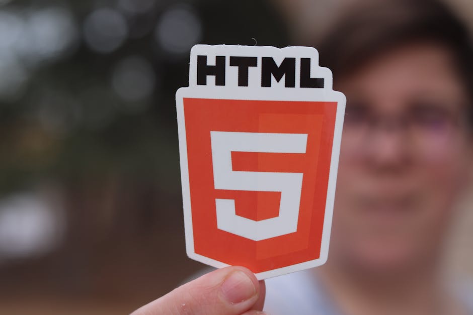 A person is holding a cut-out sign featuring the HTML5 logo, which consists of a shield shape with a prominent orange-red background and a white number '5' in the center. The top of the sign displays 